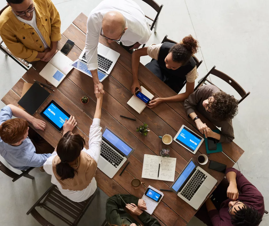 Several people around a table with various technology (laptops, tablets, and phone) in front of them, having a business meeting.