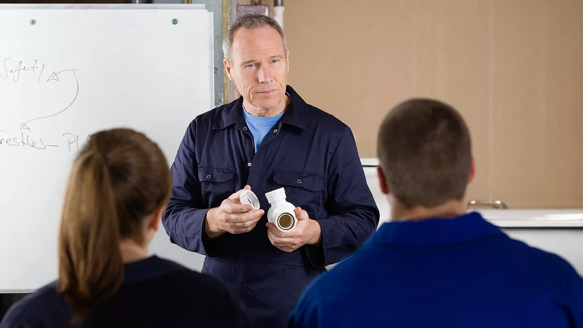 The back of a man and a woman sitting down, listening to an instructor giving plumbing lessons.