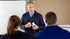 The back of a man and a woman sitting down, listening to an instructor giving plumbing lessons.