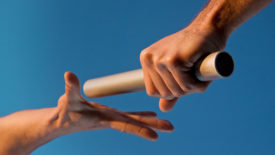 Low angle close-up of two hands passing a relay baton, with focus on the hand holding the baton, against a clear blue sky.