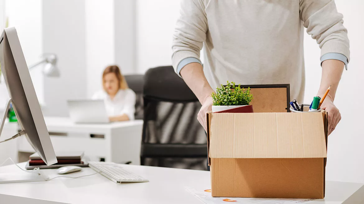 Employee packing a box and leaving the office, another employee sitting at her desk in the blurred background.