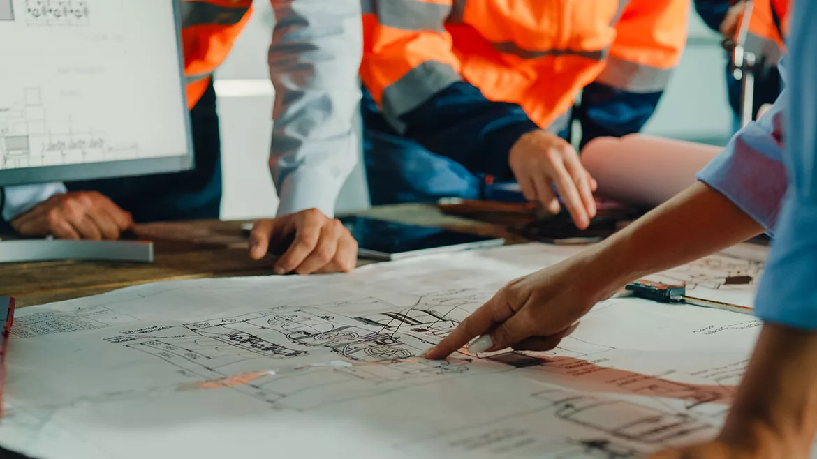 Closeup of a team of industrial engineers meeting to analyze machinery blueprints and consult on a project on a table in a manufacturing factory.