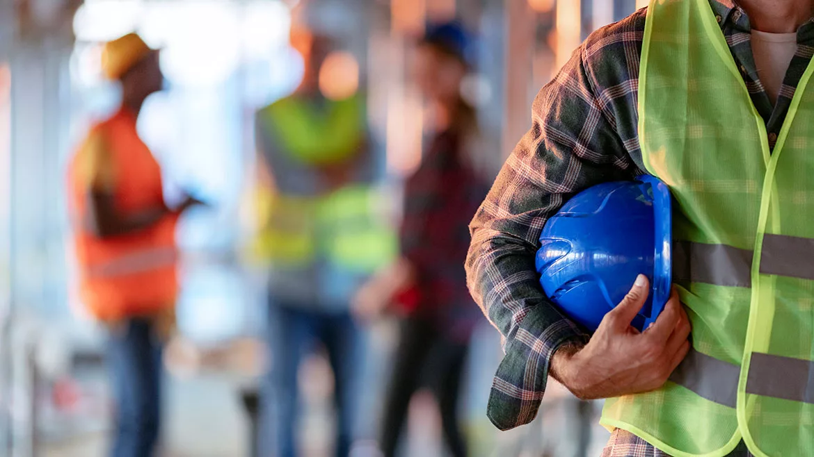 Close-up of a man holding a blue helmet. Construction worker with office and people in background. Close-up of a construction worker's hand holding a working helmet.