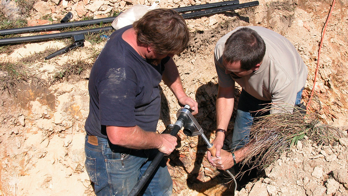 Two men fusion welding HDPE plastic by heating pipe and fitting outside surrounded by dirt.