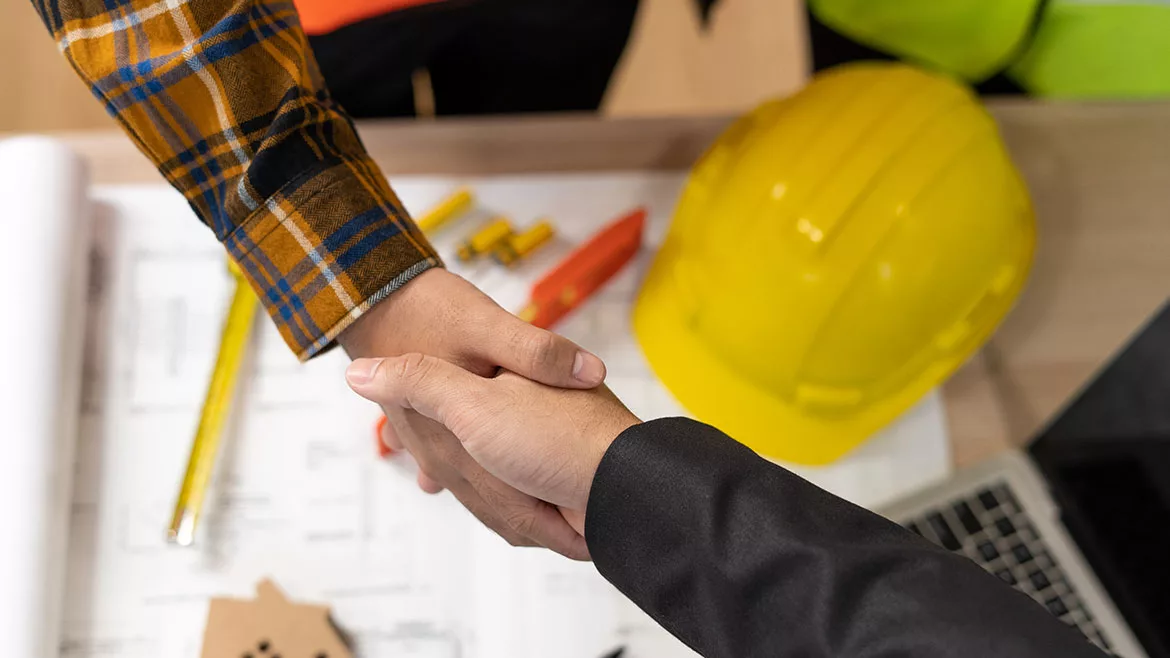 Plans and a yellow safety hat on the table while two people shake hands.