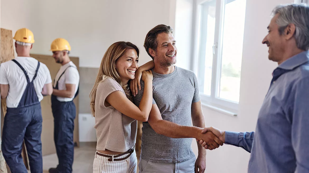 Happy couple shaking hands with their real estate agent after successful deal in the apartment. Workers wearing yellow safety hats and overalls are in the background.
