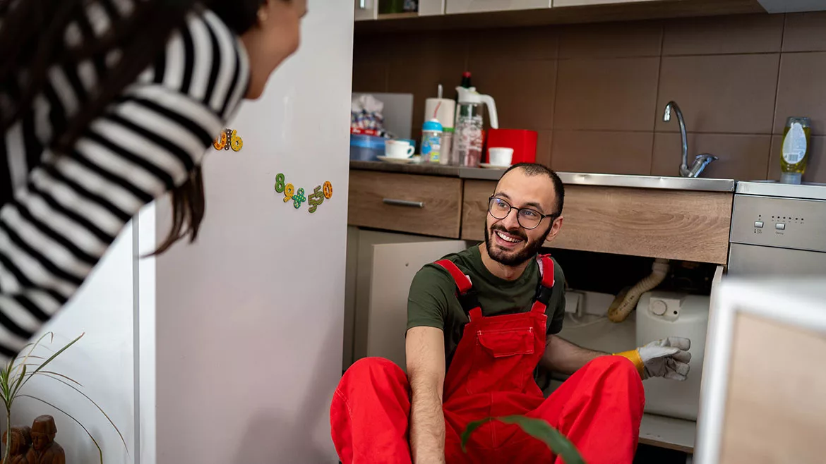 A young male plumber in red overalls is smiling and talking with a woman while working under the kitchen sink.