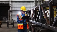 A young African-American woman working in a metal fabrication factory, wearing a hardhat, reflective vest, and safety glasses, using a digital tablet. She is standing next to a steel truss.