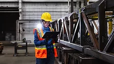 A young African-American woman working in a metal fabrication factory, wearing a hardhat, reflective vest, and safety glasses, using a digital tablet. She is standing next to a steel truss.