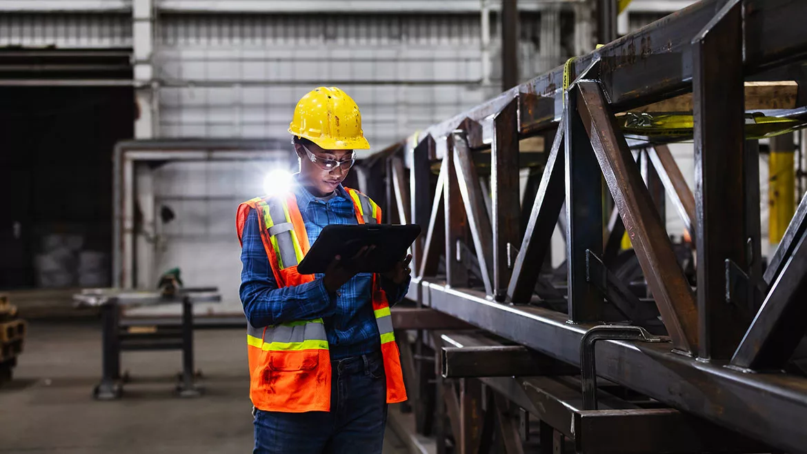 A young African-American woman working in a metal fabrication factory, wearing a hardhat, reflective vest, and safety glasses, using a digital tablet. She is standing next to a steel truss.