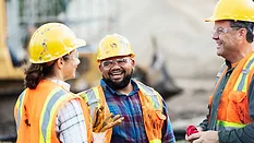 A group of three multi-ethnic workers at a construction site wearing hard hats, safety glasses and reflective clothing, smiling and conversing.