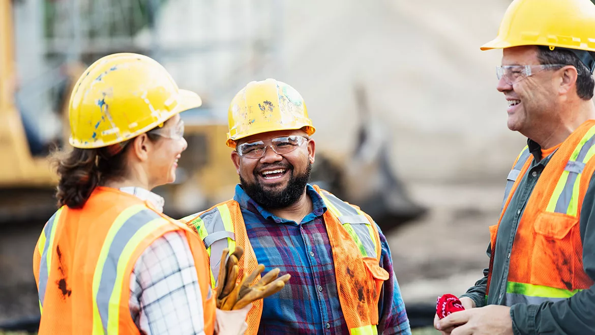 A group of three multi-ethnic workers at a construction site wearing hard hats, safety glasses and reflective clothing, smiling and conversing.
