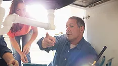 Plumbers working on pipes under sink and talking to the woman in the background.