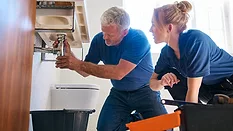 Male Plumber Fixing Leak In Domestic Bathroom Sink With Female Trainee Holding Digital Tablet.