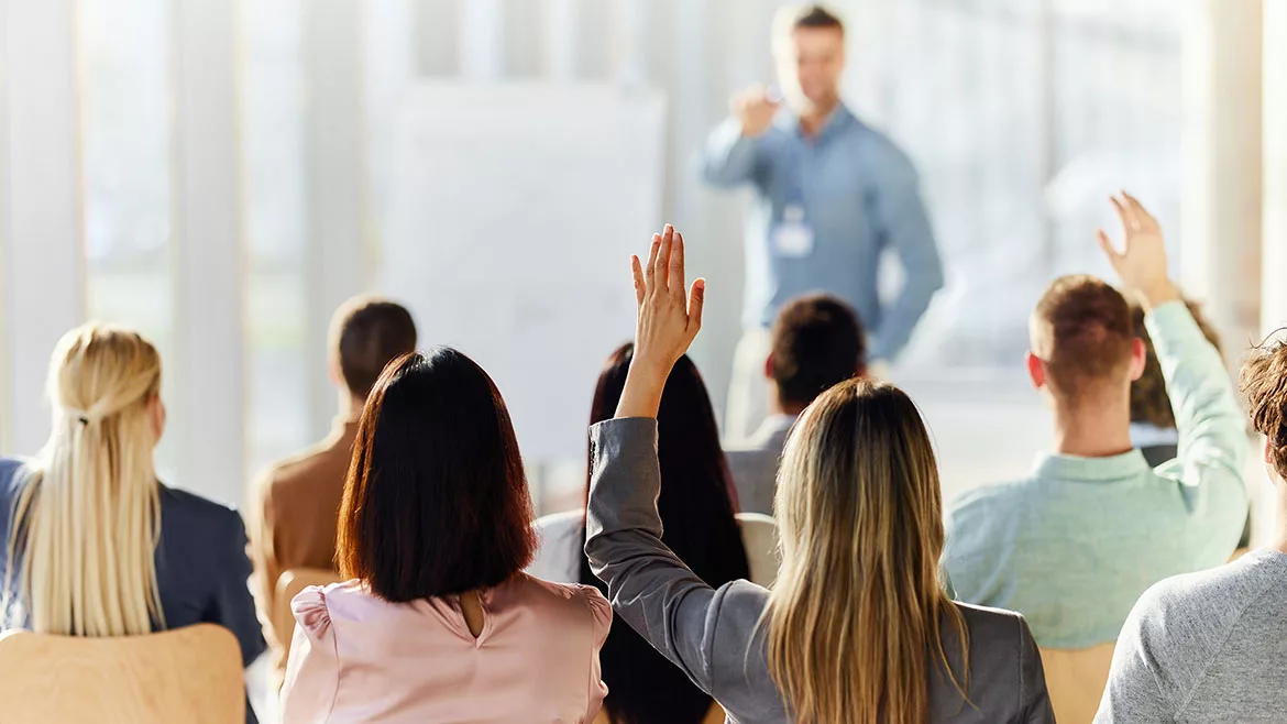 Back of seated students with raised hands in a classroom, a blurred background of the teacher pointing.