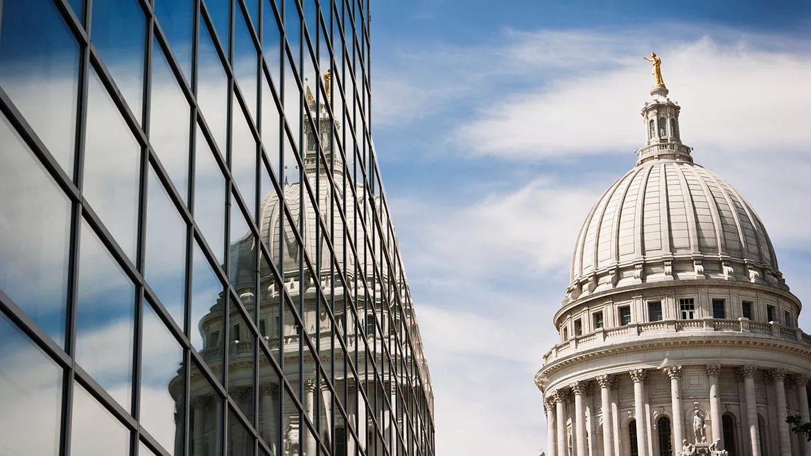 Horizontal view of the Wisconsin State Capitol Dome reflecting into the windows of a steel and glass office building on a sunny day.