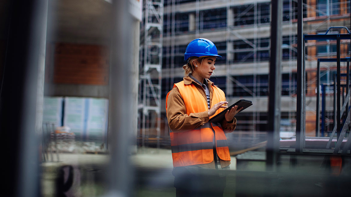 Young female worker in orange overalls and blue safety hat using her digital tablet at a construction site.