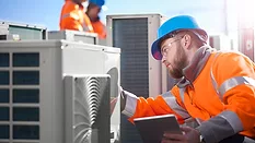 An air conditioning engineer is finishing the installation of several units on a rooftop. Two colleagues can be seen also installing units in the background. They are wearing high visibility jackets, hard hats and safety goggles.