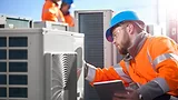An air conditioning engineer is finishing the installation of several units on a rooftop. Two colleagues can be seen also installing units in the background. They are wearing high visibility jackets, hard hats and safety goggles.