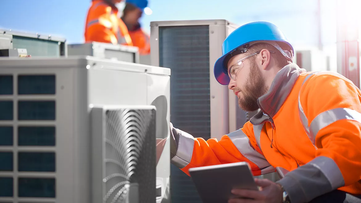 An air conditioning engineer is finishing the installation of several units on a rooftop. Two colleagues can be seen also installing units in the background. They are wearing high visibility jackets, hard hats and safety goggles.