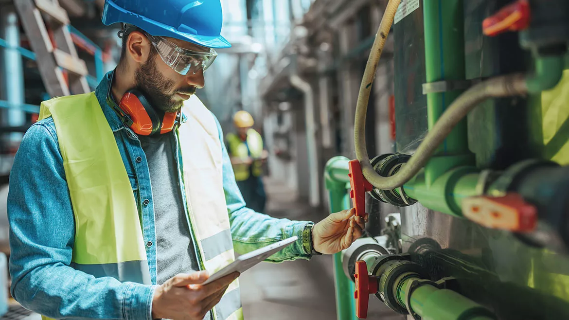 A male engineer is examining the pipe system and checking the data on his tablet.