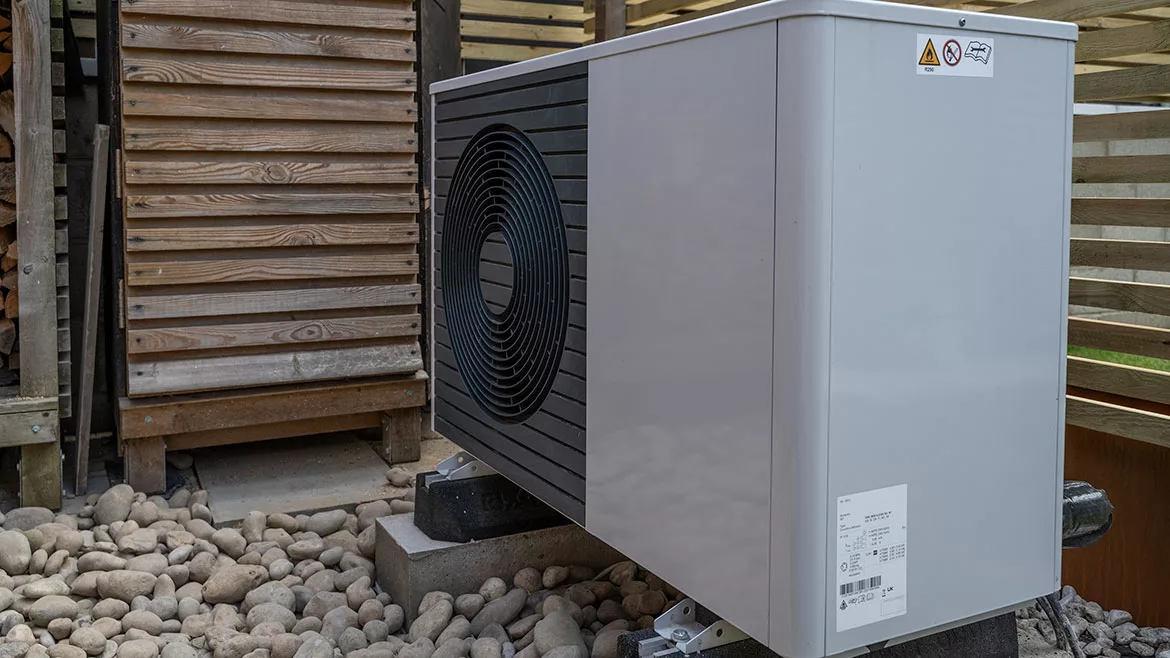 Side view of the air source heat pump outside in the garden, near wooden fence on a sunny day.