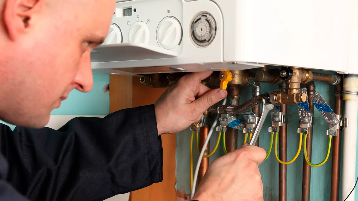 A plumber adjusting a boiler's pipes with a wrench and turning a valve.