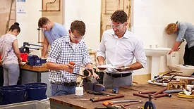 Teacher helping a college student studying plumbing. Both are wearing protective goggles while using a saw.  Other students are in the slightly blurred background.