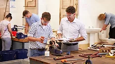 Teacher helping a college student studying plumbing. Both are wearing protective goggles while using a saw.  Other students are in the slightly blurred background.