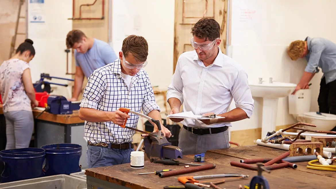 Teacher helping a college student studying plumbing. Both are wearing protective goggles while using a saw. Other students are in the slightly blurred background.