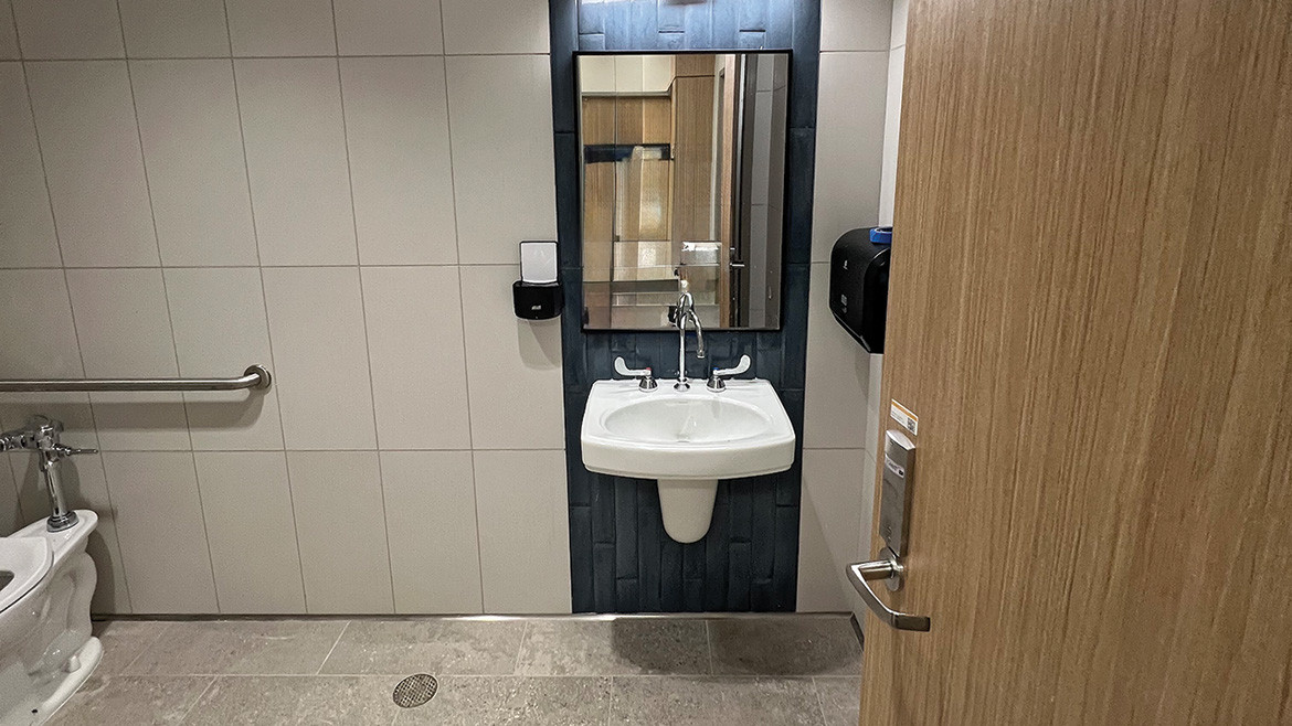 A bathroom image taken from the door showing the sink and mirro, a cropped image of the toilet and a drain in the middle of the floor.