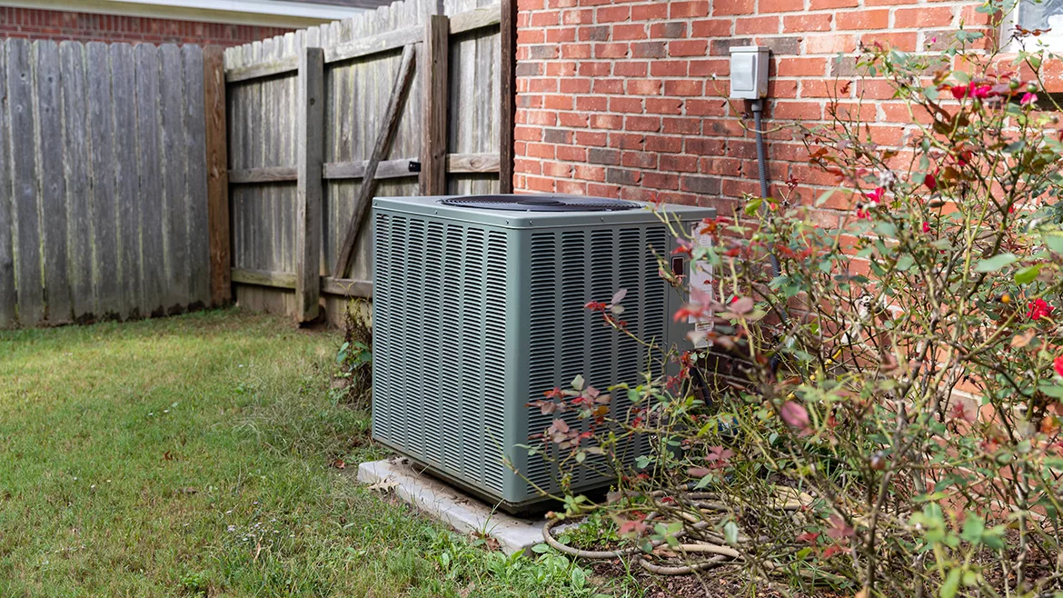 Air conditioner condenser unit sitting next to brick home with fence.