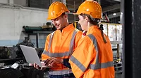 Two engineers are wearing orange safety vests and hard hats and are looking at a laptop at a locomotive service station.