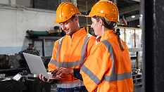 Two engineers are wearing orange safety vests and hard hats and are looking at a laptop at a locomotive service station.