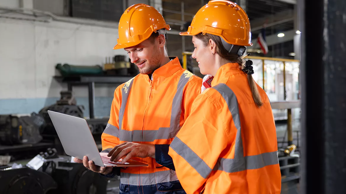 Two engineers are wearing orange safety vests and hard hats and are looking at a laptop at a locomotive service station.