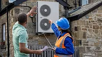 An air-source heat pump is being installed on a home in North East England for sustainability. A female construction worker in a hard hat and reflective jacket points to the heat pump while speaking to a man holding papers and gesturing.