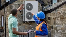 An air-source heat pump is being installed on a home in North East England for sustainability. A female construction worker in a hard hat and reflective jacket points to the heat pump while speaking to a man holding papers and gesturing.