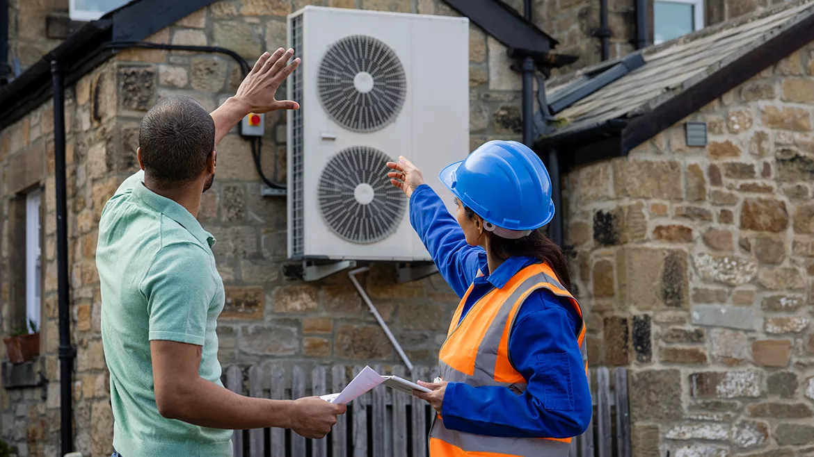 An air-source heat pump is being installed on a home in North East England for sustainability. A female construction worker in a hard hat and reflective jacket points to the heat pump while speaking to a man holding papers and gesturing.