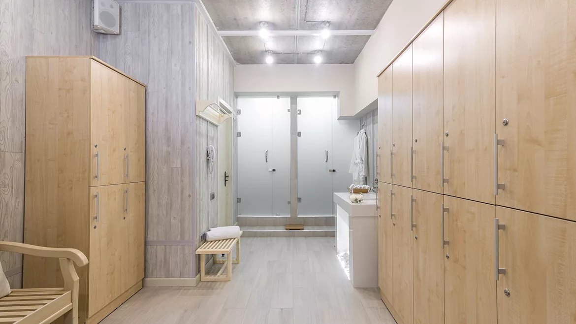 Feature image of a locker room with wooden lockers, a robe, hand towels in a basket and two showers in the back.