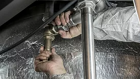 Plumber's hands with dirty sleeves, gripping pipes to turn it, a silver thermal material in the background.