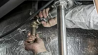 Plumber's hands with dirty sleeves, gripping pipes to turn it, a silver thermal material in the background.
