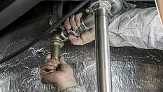 Plumber's hands with dirty sleeves, gripping pipes to turn it, a silver thermal material in the background.